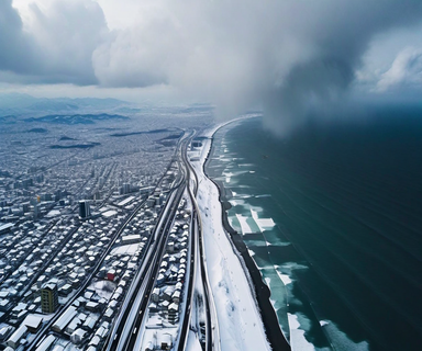 Aerial view of Japan's coast during severe winter storm showing heavy snow clouds from the Japan Sea moving over snow-covered land with transportation routes visible below