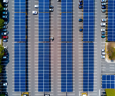 Aerial view of a public parking lot with solar panel canopies installed over parking spaces, showing organized rows of blue photovoltaic panels generating renewable energy in an urban setting